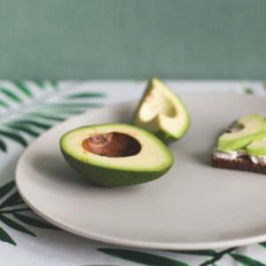 On a plate is a ripe avocado and a sandwich of cereal bread, soft cheese and slices of avocado